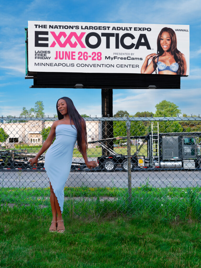 Vanniall in a blue dress on a lawn in front of a large EXXXotica Billboard featuring her image!