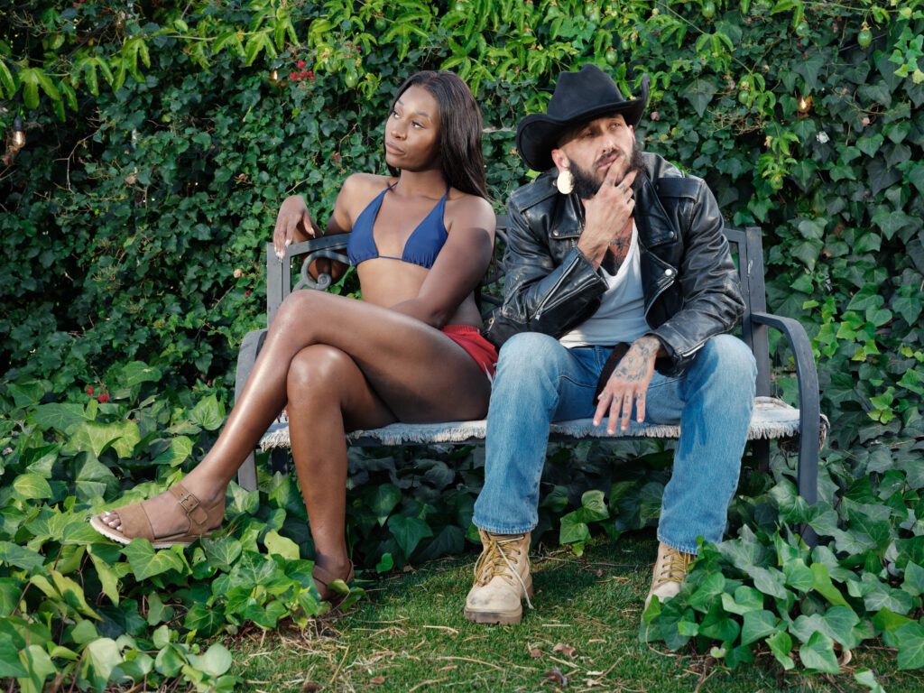 Vanniall (left) and Viktor Belmont (right) on the set of "The Cowboy and the Rose," seated on a bench against green foliage.